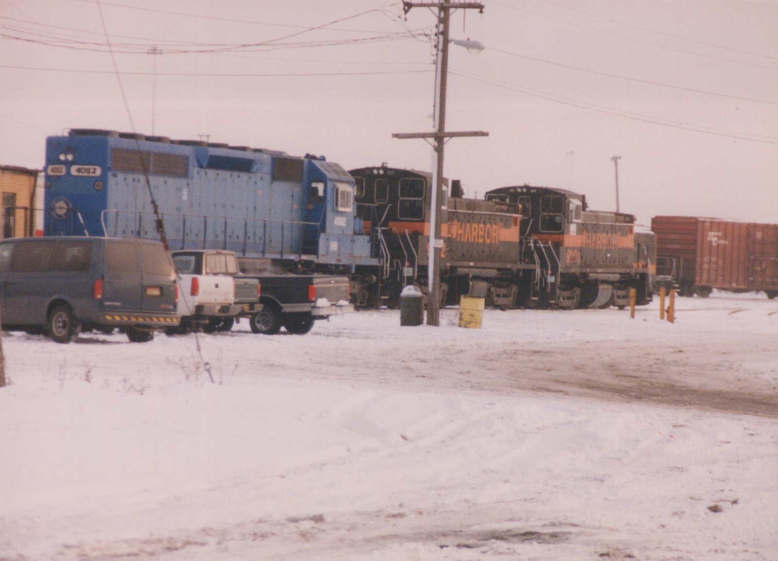 A blue diesel locomotive numbered 4002 and two black-and-orange switchers lettered HARBOR stand in a snow-covered yard, with a van and two pickup trucks parked in front and a red boxcar at right.