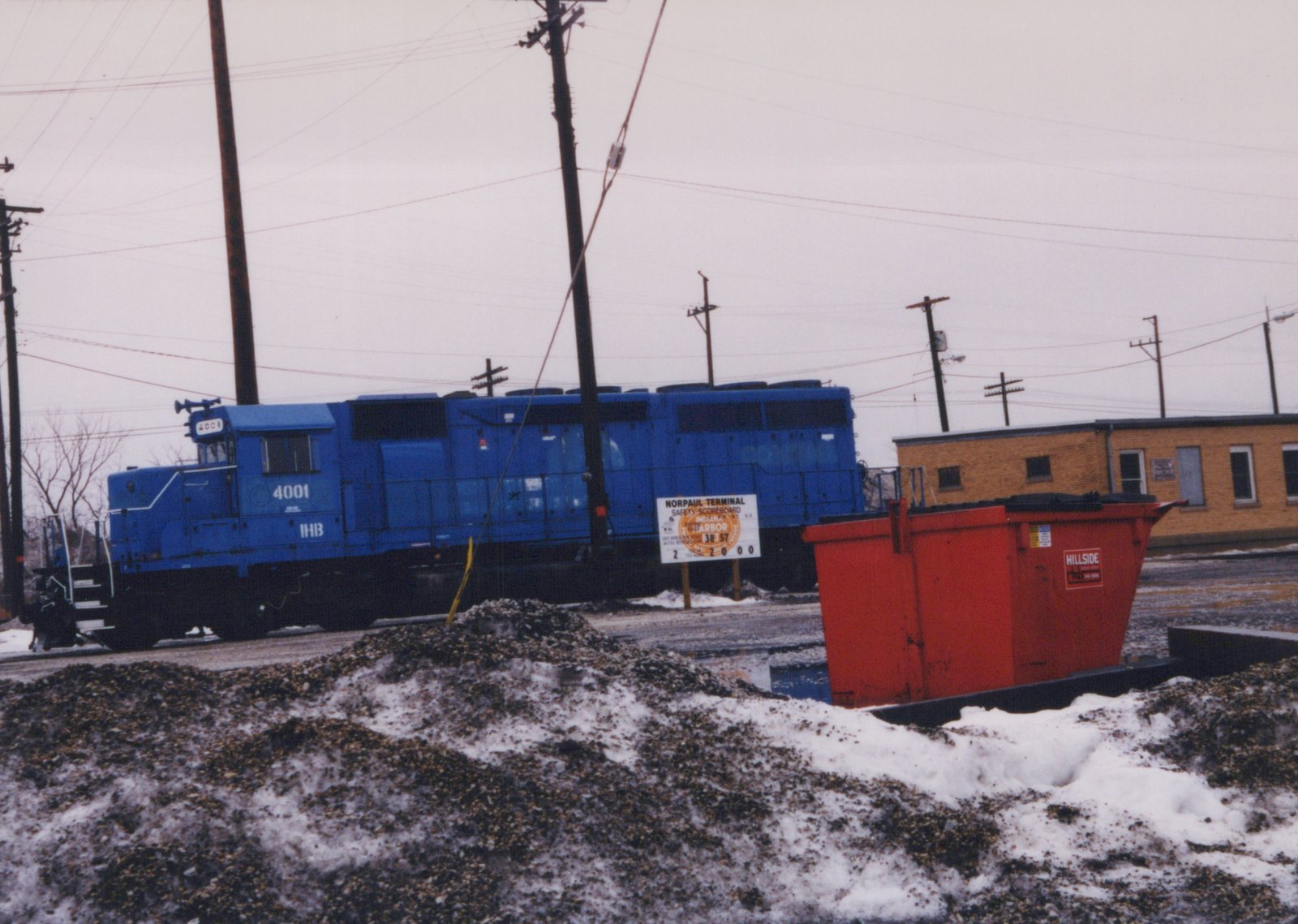 A blue diesel locomotive numbered 4001 and lettered IHB stands on a track behind a pile of gravel and snow, with a safety sign, a red dumpster and a yellow brick building at right.