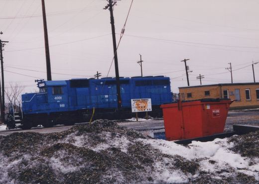 A blue diesel locomotive numbered 4001 and lettered IHB stands on a track behind a pile of gravel and snow, with a safety sign, a red dumpster and a yellow brick building at right.