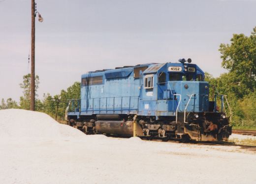 A blue six-axle diesel locomotive numbered 4002 stands on a track beside a large pile of white gravel, with trees and a wooden lamp pole behind it.