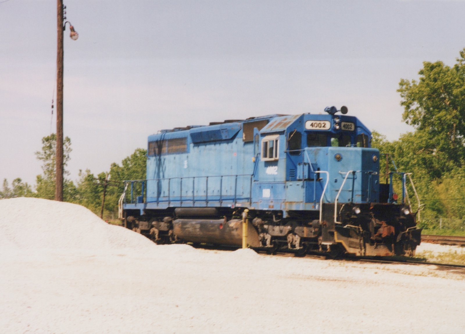 A blue six-axle diesel locomotive numbered 4002 stands on a track beside a large pile of white gravel, with trees and a wooden lamp pole behind it.