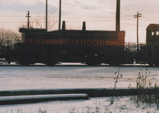 Side view of a black locomotive with no cab, lettered HARBOR in orange, coupled to the cab of another switcher at right, with snow in the foreground and utility poles against a pale sky.