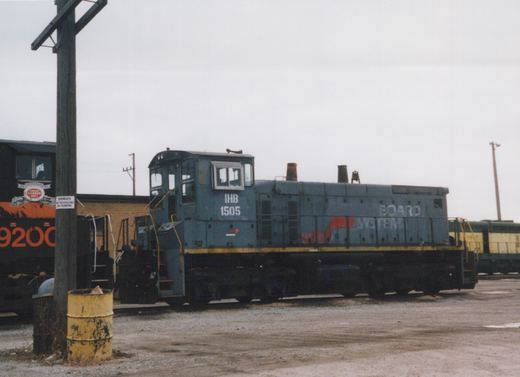 A gray diesel switcher lettered IHB 1505, with BOARD SYSTEM still on its hood, sits on gravel in a rail yard under an overcast sky. A black and orange locomotive stands at left, and a yellow and green locomotive is at right.
