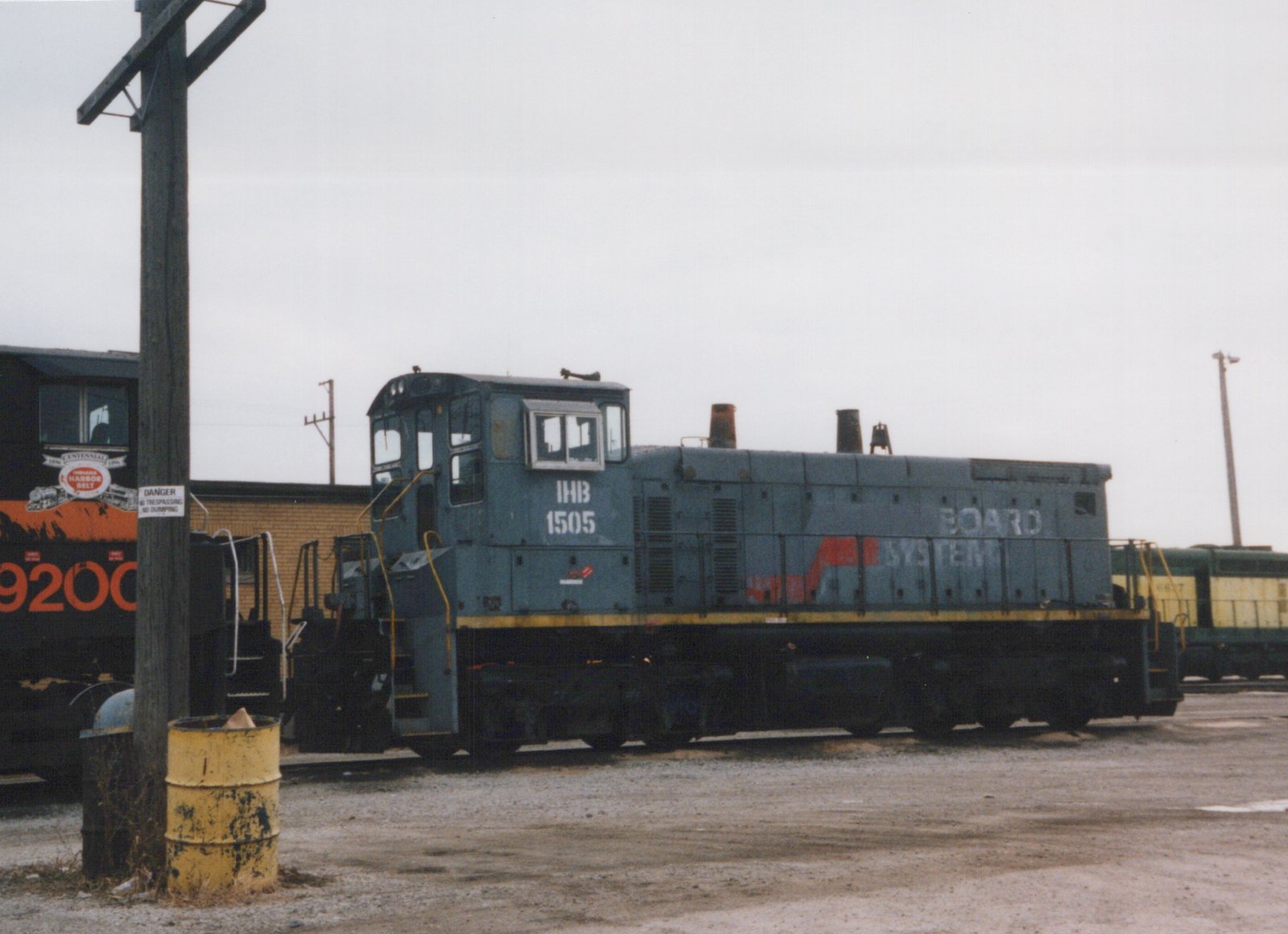 A gray diesel switcher lettered IHB 1505, with BOARD SYSTEM still on its hood, sits on gravel in a rail yard under an overcast sky. A black and orange locomotive stands at left, and a yellow and green locomotive is at right.
