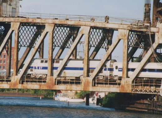 A silver locomotive with a blue stripe and the number 27, followed by a passenger car lettered Coach, crosses a rusty steel truss bridge above a river with moored boats.