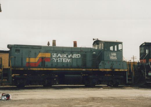 Side view of a gray end-cab switcher with a yellow and red stripe and SEABOARD SYSTEM lettering on its hood, with IHB 1505 stenciled on the cab. A black locomotive stands behind it under an overcast sky.