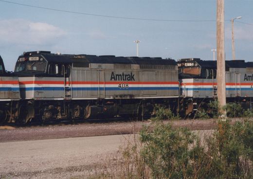 A line of silver Amtrak diesel locomotives with red, white and blue stripes stands in a yard behind gravel and shrubs, with number 408 in the center.