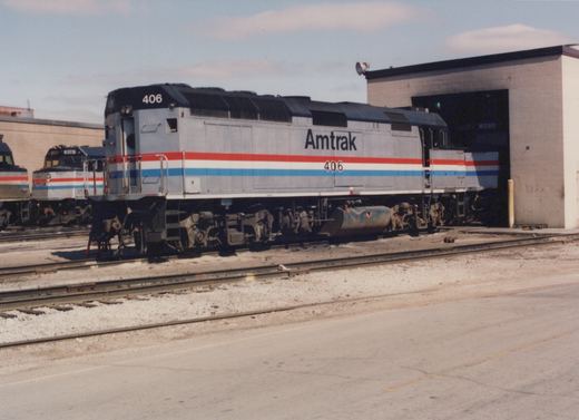 A silver locomotive with red, white and blue stripes, lettered Amtrak 406, stands on a yard track. Its far end reaches into the open door of a concrete-block shop building, and two more Amtrak locomotives sit to the left.