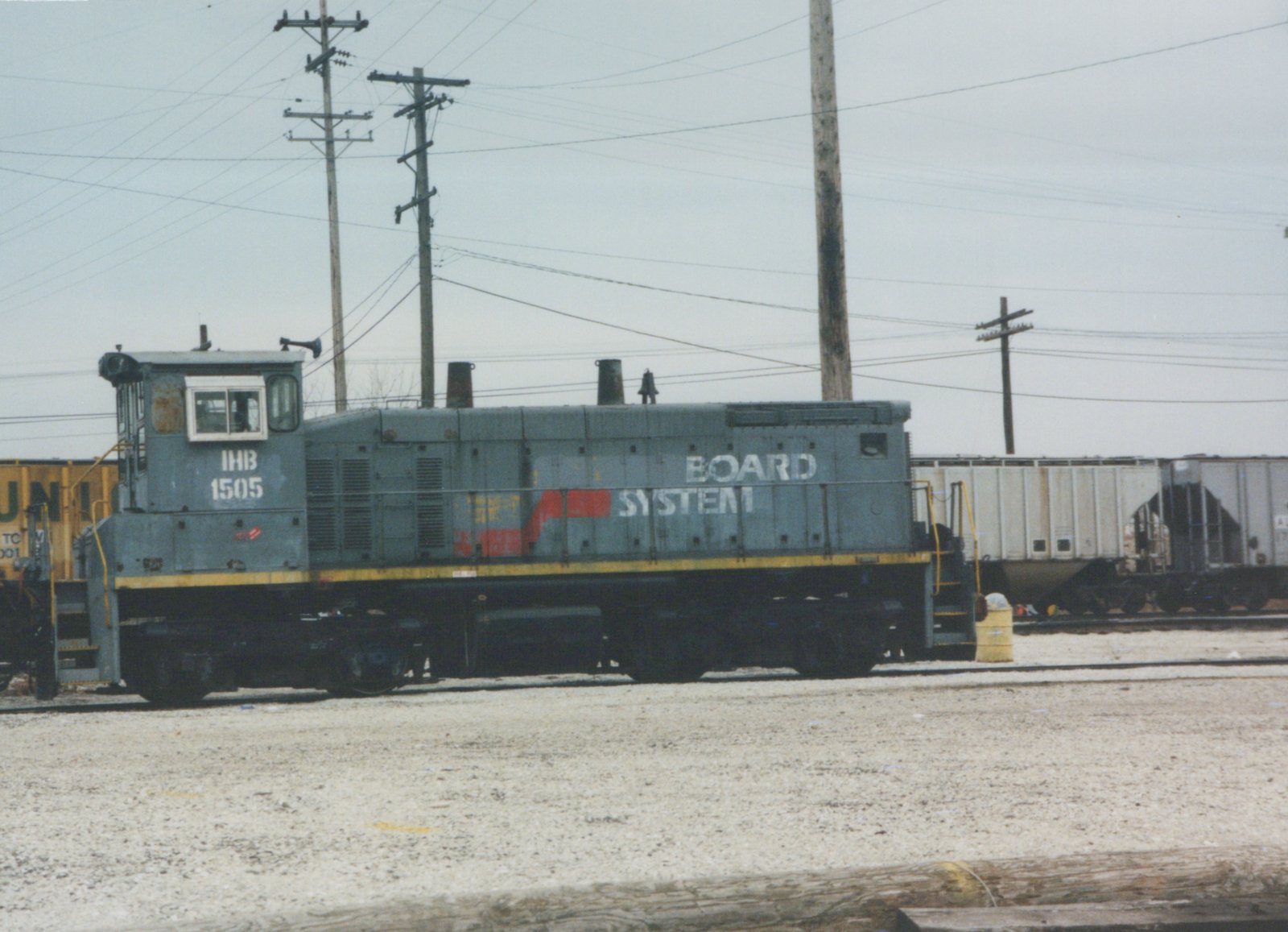 A grey end-cab diesel switcher lettered IHB 1505 on its cab, with BOARD SYSTEM still on its hood, stands on a gravel yard track under utility poles, with covered hoppers behind it.