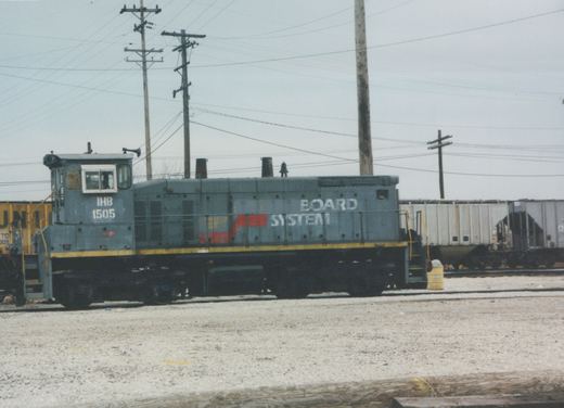 A grey end-cab diesel switcher lettered IHB 1505 on its cab, with BOARD SYSTEM still on its hood, stands on a gravel yard track under utility poles, with covered hoppers behind it.