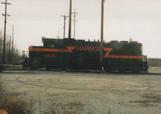 Side view of a black diesel locomotive with orange stripes and HARBOR lettering, number 2920 on the cab, standing on a track beyond a gravel lot under an overcast sky, with utility poles around it.