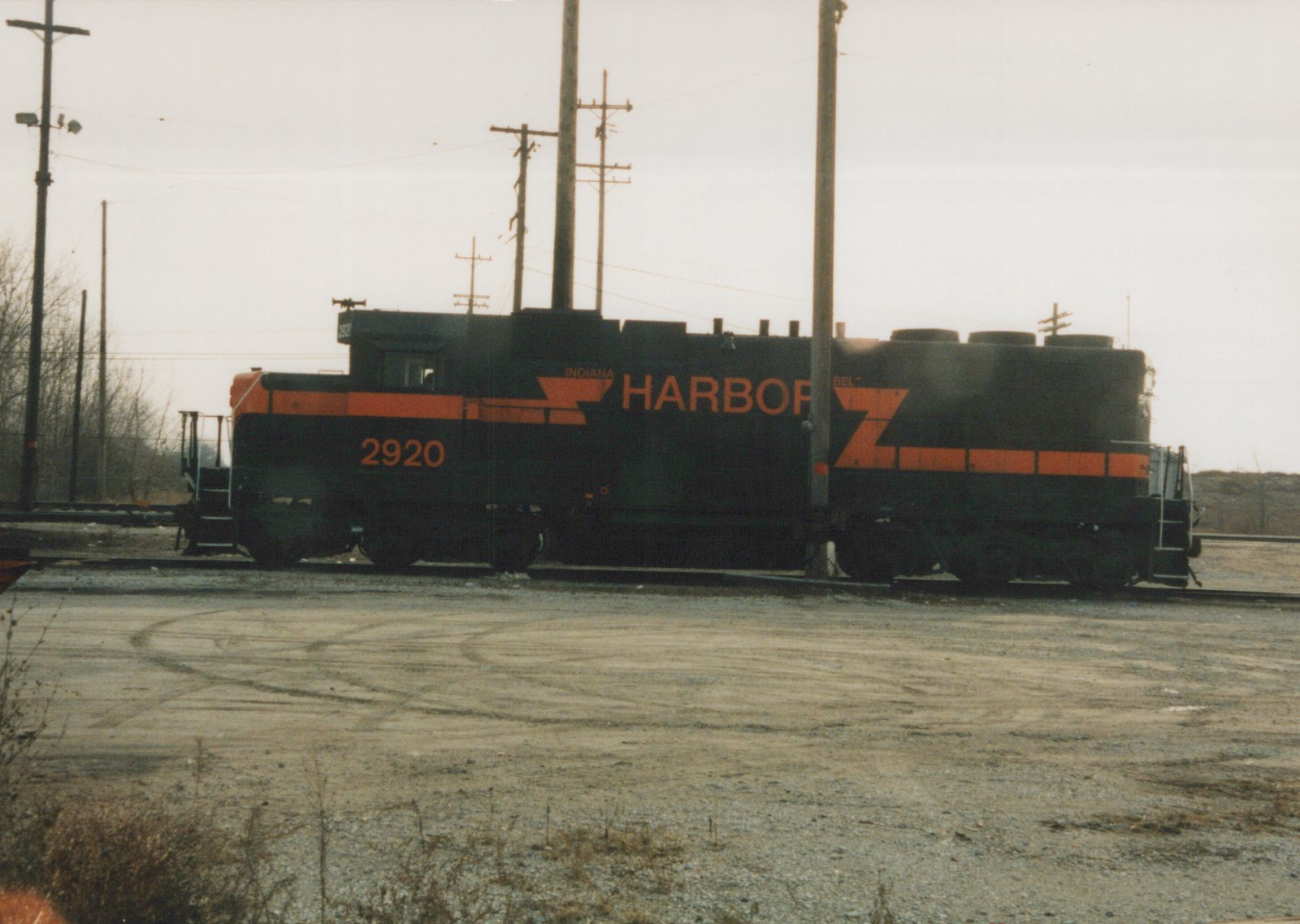Side view of a black diesel locomotive with orange stripes and HARBOR lettering, number 2920 on the cab, standing on a track beyond a gravel lot under an overcast sky, with utility poles around it.
