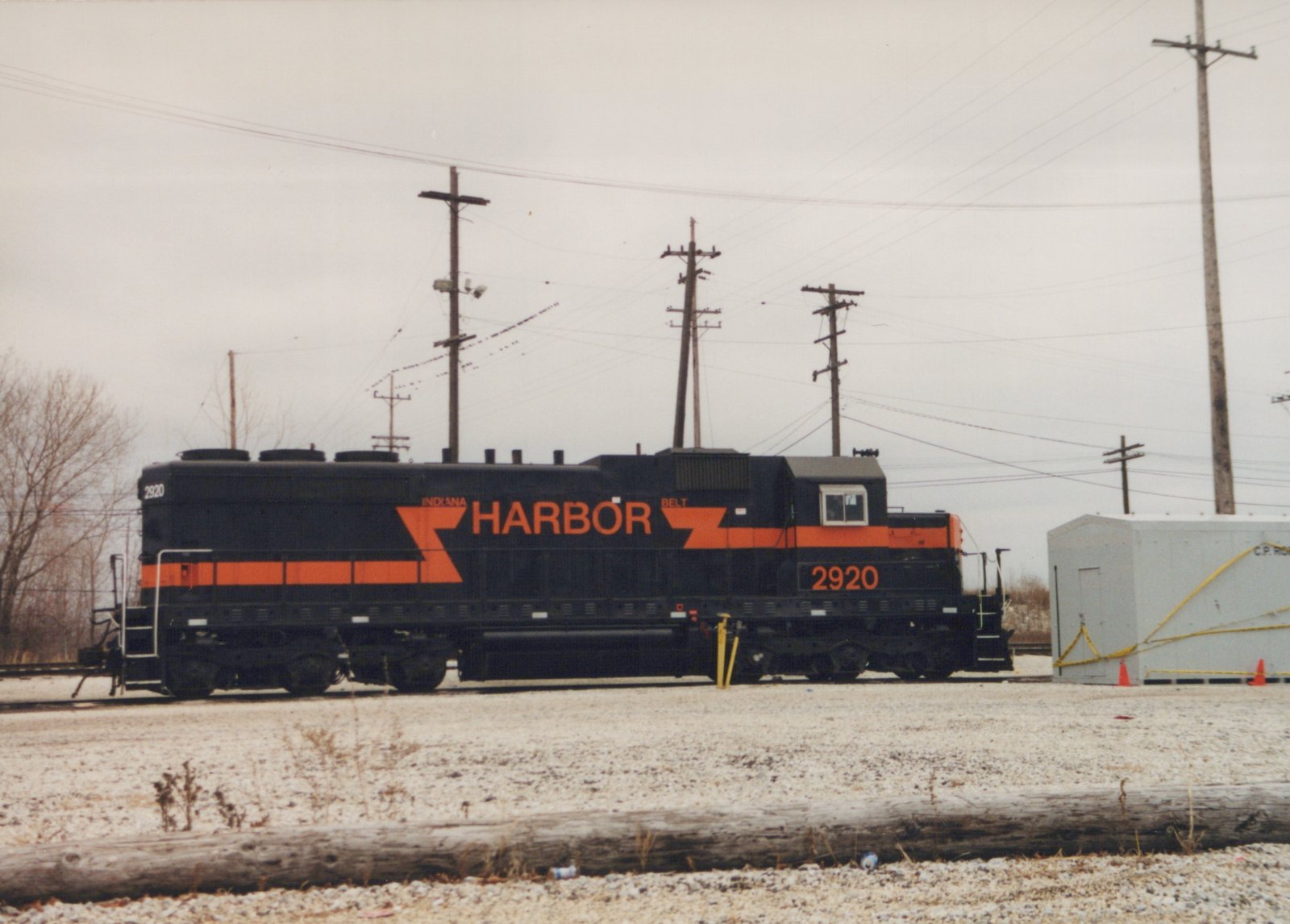 Side view of a black locomotive with an orange stripe, orange lightning-bolt graphics and HARBOR in large orange letters, standing on a lightly snow-dusted track beside a grey equipment shelter, with utility poles behind.