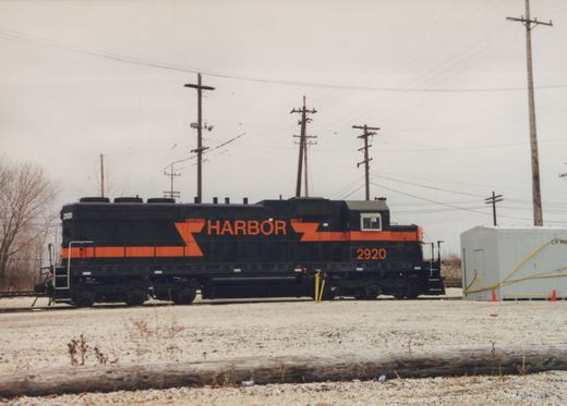 Side view of a black locomotive with an orange stripe, orange lightning-bolt graphics and HARBOR in large orange letters, standing on a lightly snow-dusted track beside a grey equipment shelter, with utility poles behind.