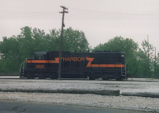 A black diesel locomotive with orange stripes and large orange HARBOR lettering, number 2920, standing on gravel-ballasted tracks under an overcast sky, with green trees and a leaning utility pole behind it.