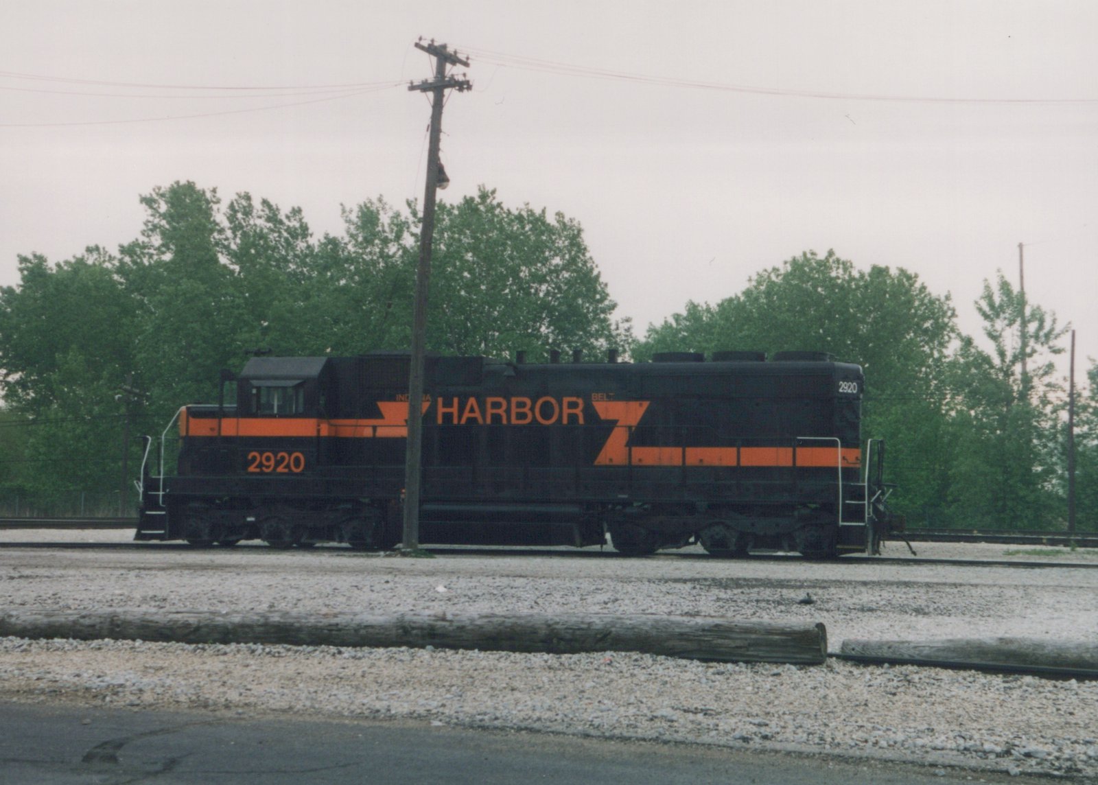 A black diesel locomotive with orange stripes and large orange HARBOR lettering, number 2920, standing on gravel-ballasted tracks under an overcast sky, with green trees and a leaning utility pole behind it.