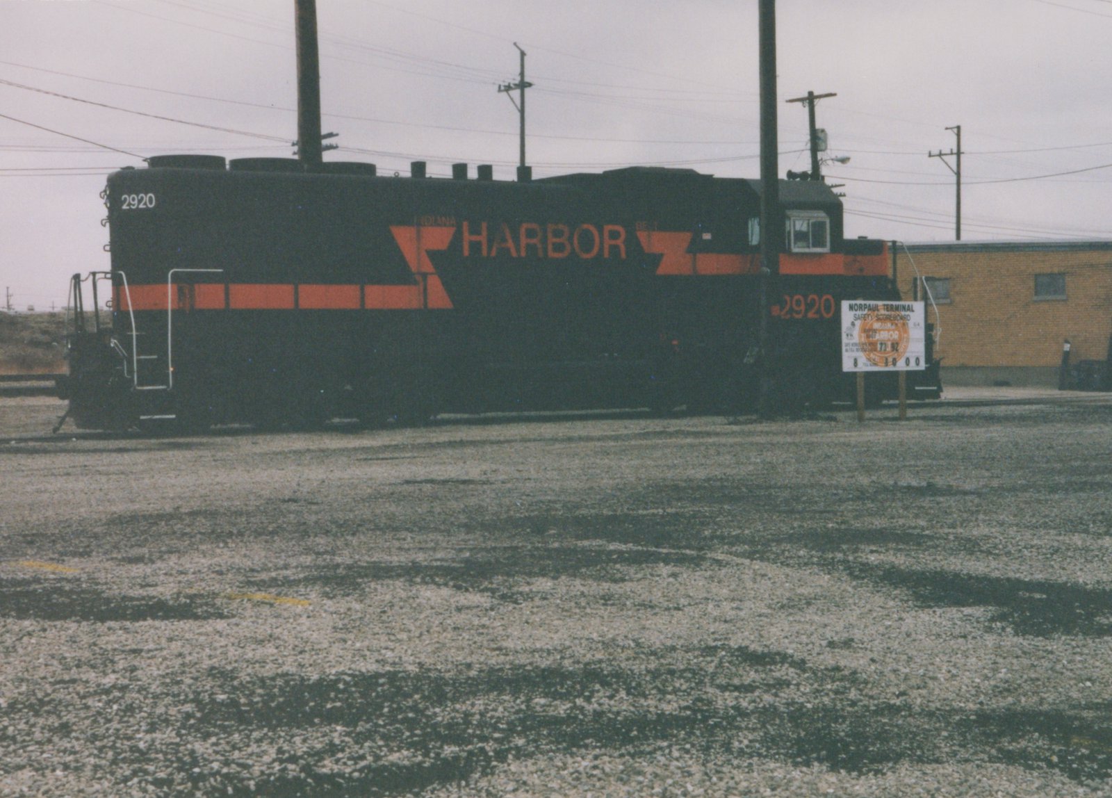A black diesel locomotive with orange stripes and HARBOR lettering, number 2920, stands on a gravel yard beside a Norpaul Terminal safety scoreboard sign under an overcast sky.
