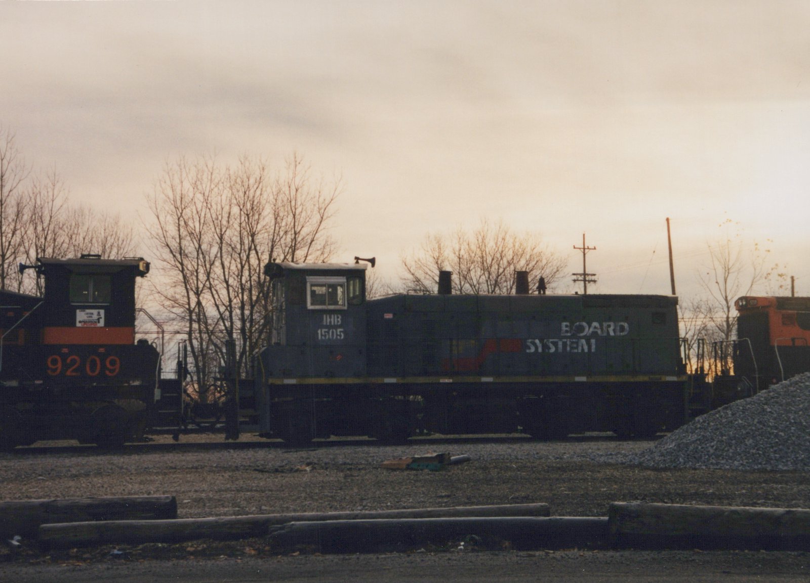 A gray end-cab diesel switcher lettered IHB 1505, with BOARD SYSTEM on its hood, stands between a black and orange locomotive numbered 9209 and an orange locomotive under an overcast evening sky.