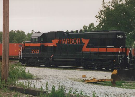 A black diesel locomotive with an orange stripe and large orange HARBOR lettering stands on a track beside gravel and weeds, with orange shipping containers and trees behind it under an overcast sky.