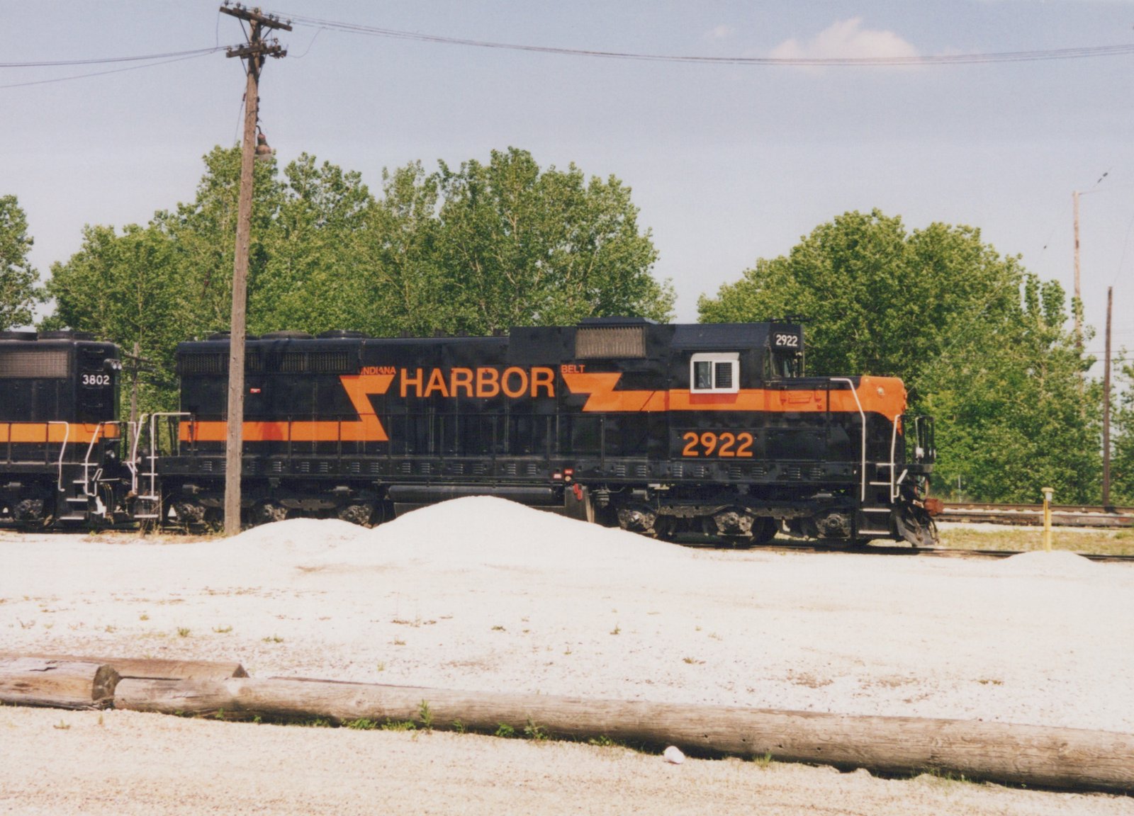 Side view of a black diesel locomotive with an orange stripe and the words INDIANA HARBOR BELT, numbered 2922, coupled to a second black and orange locomotive numbered 3802, behind a gravel lot with piles of ballast and logs, with trees and a utility pole beyond.