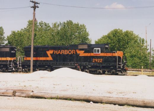 Side view of a black diesel locomotive with an orange stripe and the words INDIANA HARBOR BELT, numbered 2922, coupled to a second black and orange locomotive numbered 3802, behind a gravel lot with piles of ballast and logs, with trees and a utility pole beyond.