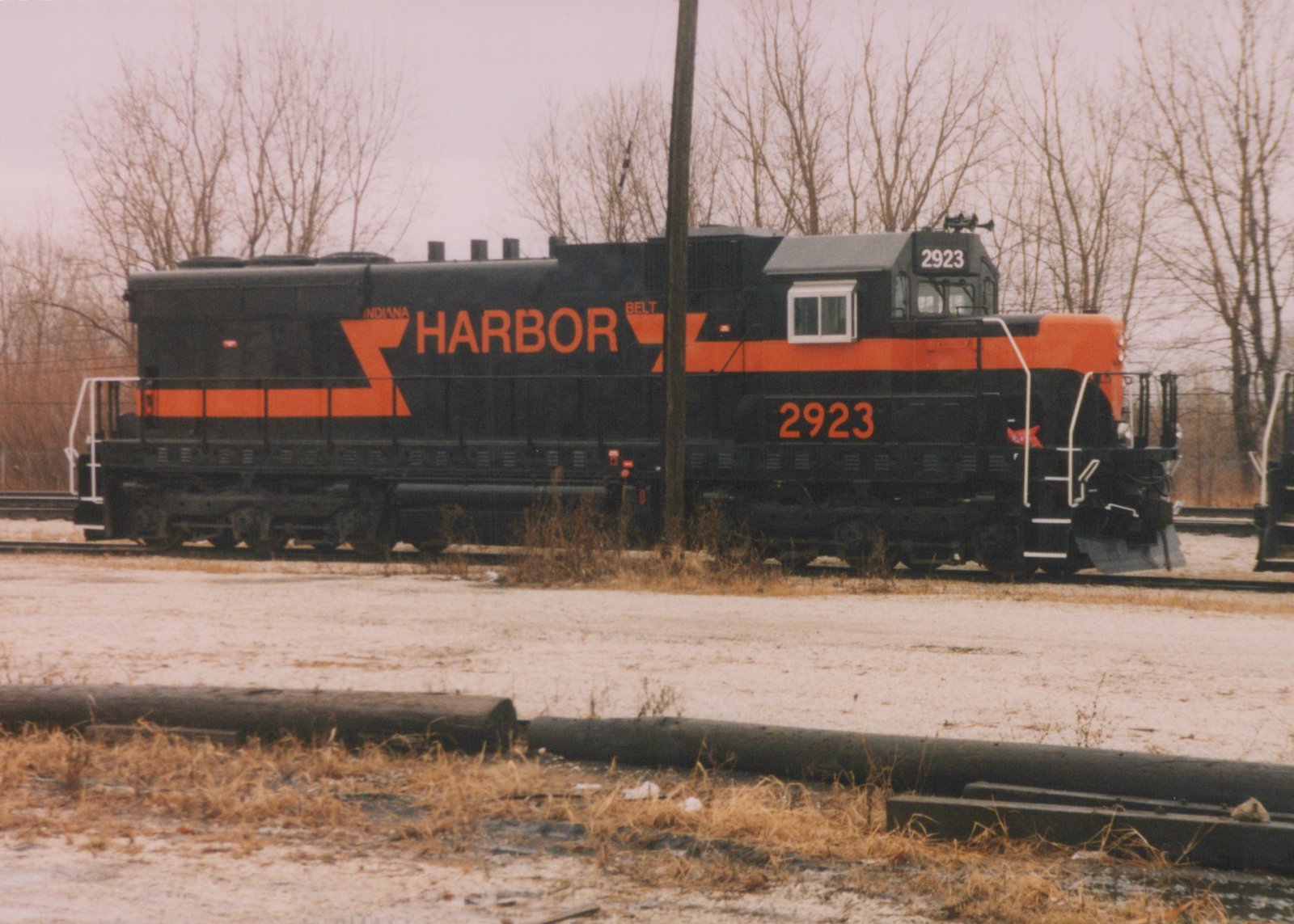 A black diesel locomotive with orange stripes, HARBOR lettering and the number 2923 stands on a yard track behind a snowy, weedy lot with bare trees behind it.