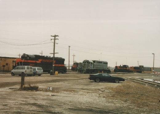 A black and orange locomotive lettered HARBOR and numbered 2923 stands behind two parked minivans in a gravel yard lot. A gray locomotive, a red-nosed SP unit and two black and orange switchers are farther back, and a dark sports car is parked in the foreground.