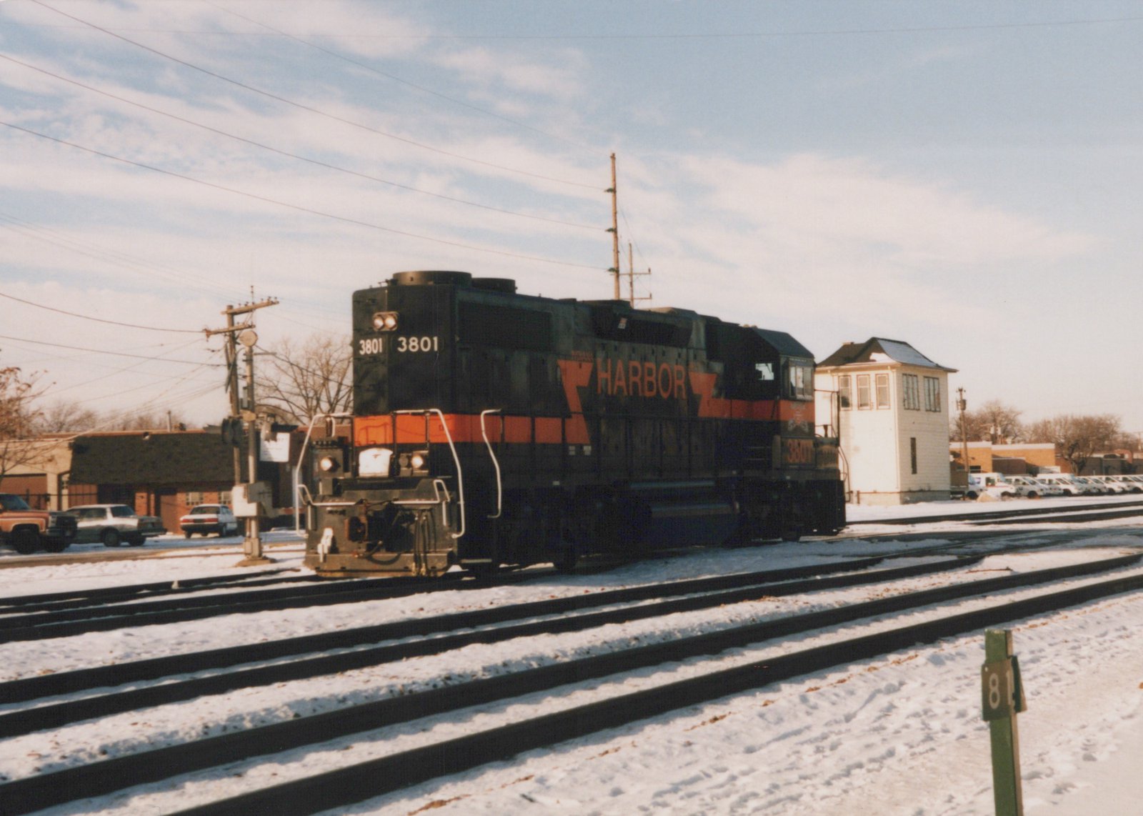 A black and orange diesel locomotive lettered HARBOR, numbered 3801, sits on snow-covered tracks next to a white two-story tower under a partly cloudy sky.