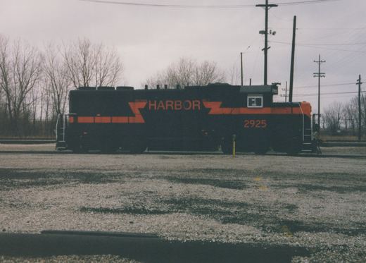 A black diesel locomotive with orange stripes and the word HARBOR on its side, number 2925 on the cab, stands on a yard track across a gravel lot with bare trees and utility poles behind it.