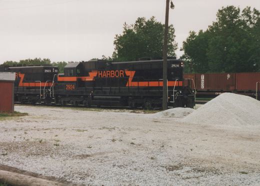 Two black diesel locomotives with orange stripes and large orange HARBOR lettering stand on a track behind a gravel lot, with red containers on a freight car and trees behind them under an overcast sky.
