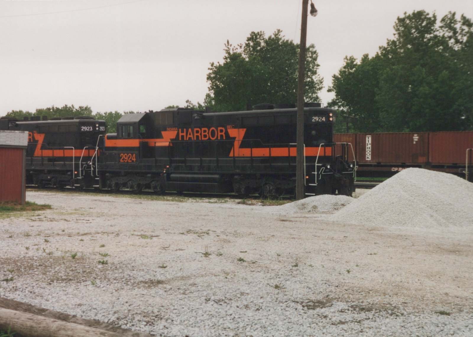 Two black diesel locomotives with orange stripes and large orange HARBOR lettering stand on a track behind a gravel lot, with red containers on a freight car and trees behind them under an overcast sky.