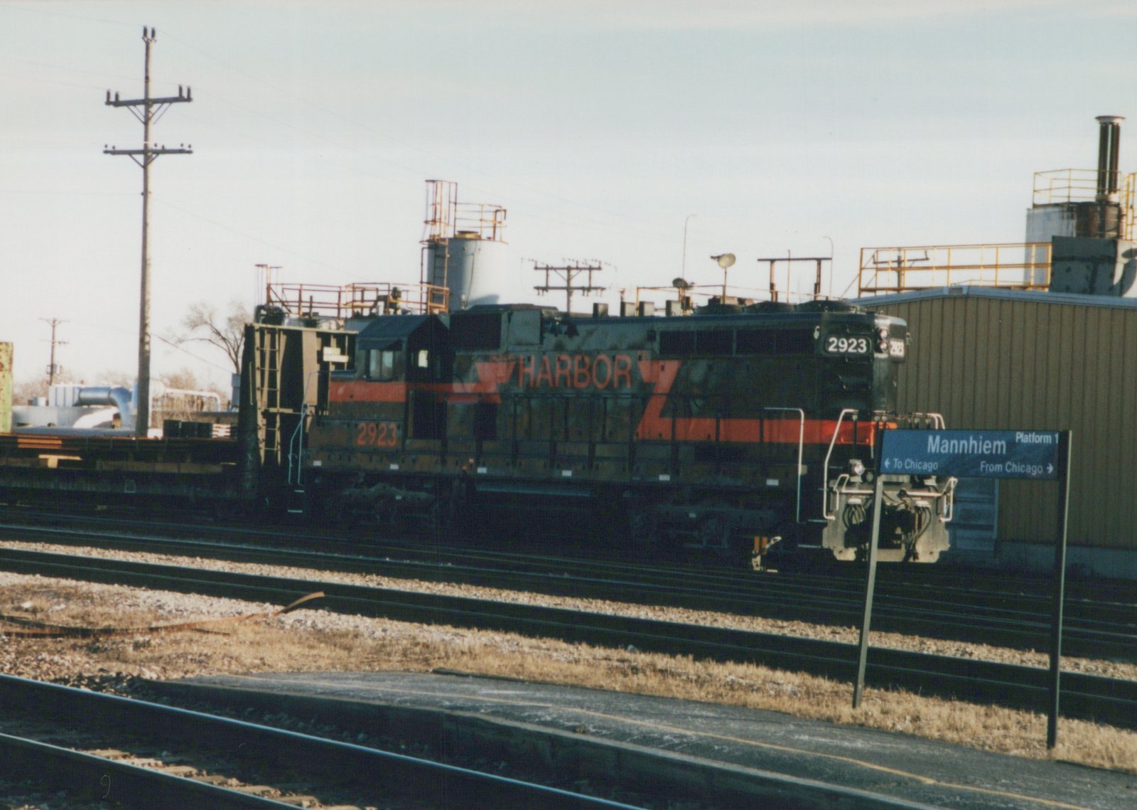 A black diesel locomotive with orange stripes and HARBOR lettering stands on a yard track beyond a station platform with a blue sign reading Mannhiem, Platform 1.