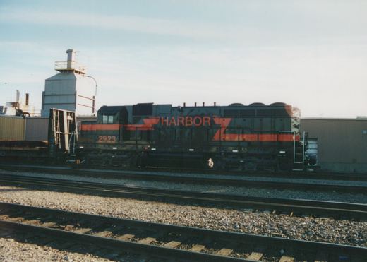 Side view of a black diesel locomotive with orange stripes and HARBOR lettering, coupled to a bulkhead flatcar on a track across several rail lines from the camera, with industrial buildings behind.