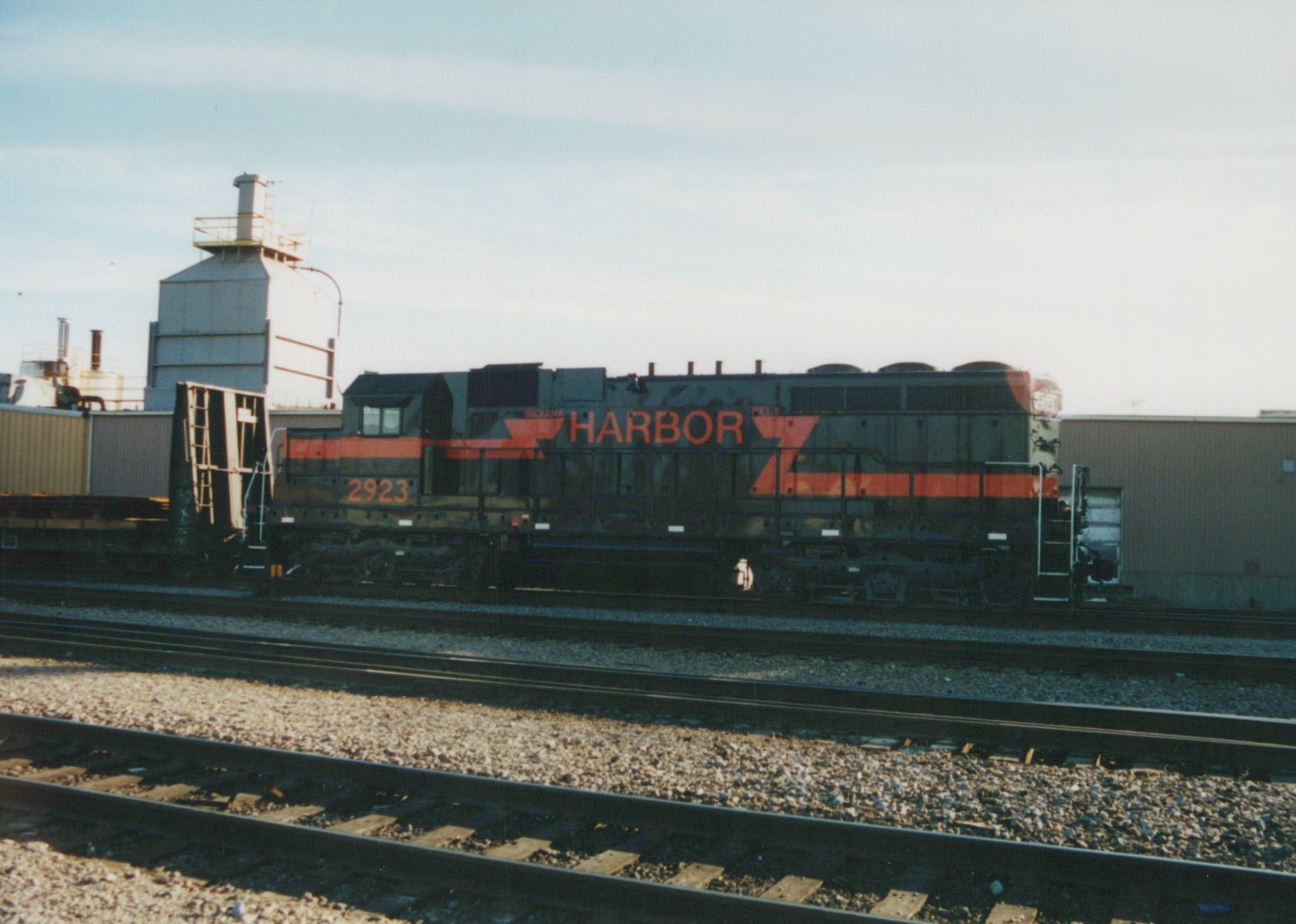 Side view of a black diesel locomotive with orange stripes and HARBOR lettering, coupled to a bulkhead flatcar on a track across several rail lines from the camera, with industrial buildings behind.