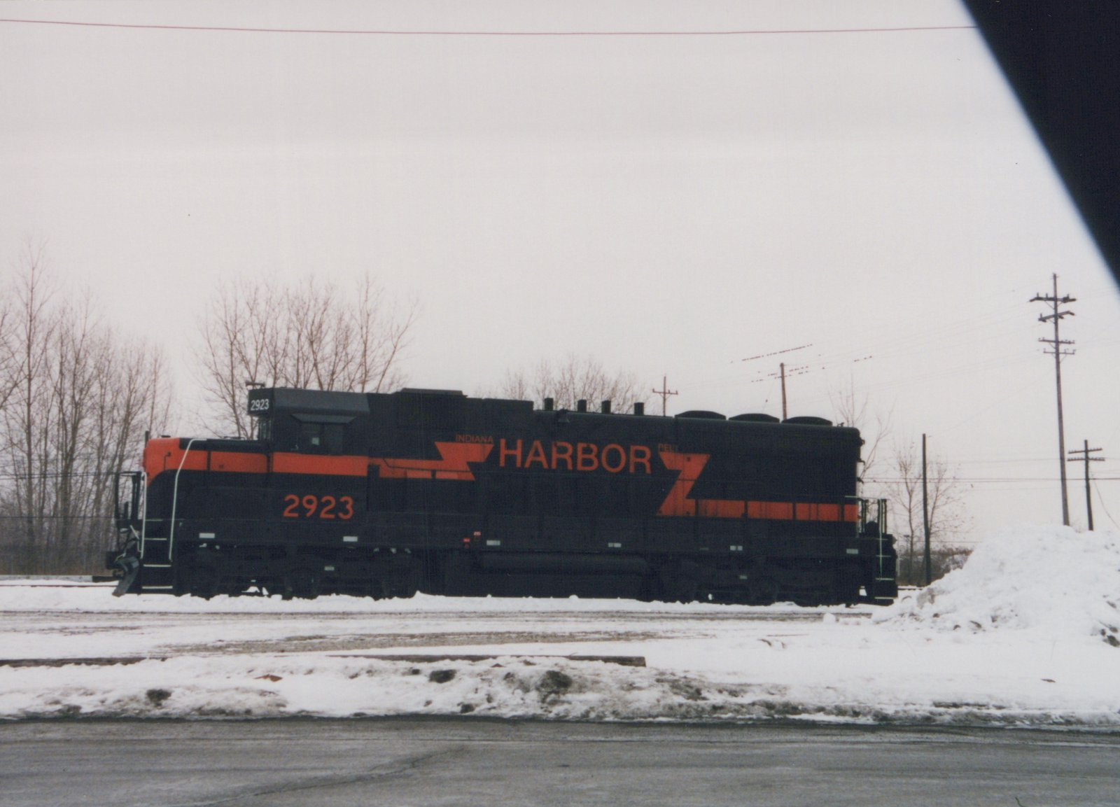 Side view of a black locomotive with an orange nose and stripe, lettered HARBOR and numbered 2923, on a snow-covered track under an overcast sky.