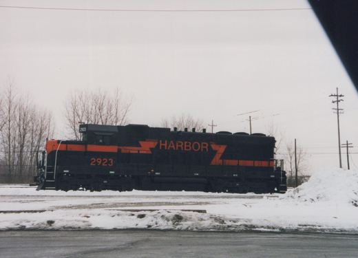 Side view of a black locomotive with an orange nose and stripe, lettered HARBOR and numbered 2923, on a snow-covered track under an overcast sky.