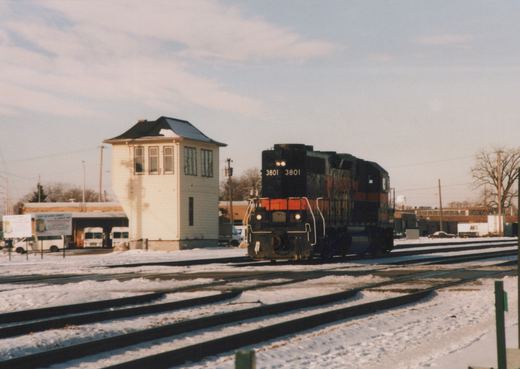A black and orange diesel locomotive numbered 3801 moves on snowy tracks beside a white two-story interlocking tower under a partly cloudy sky.