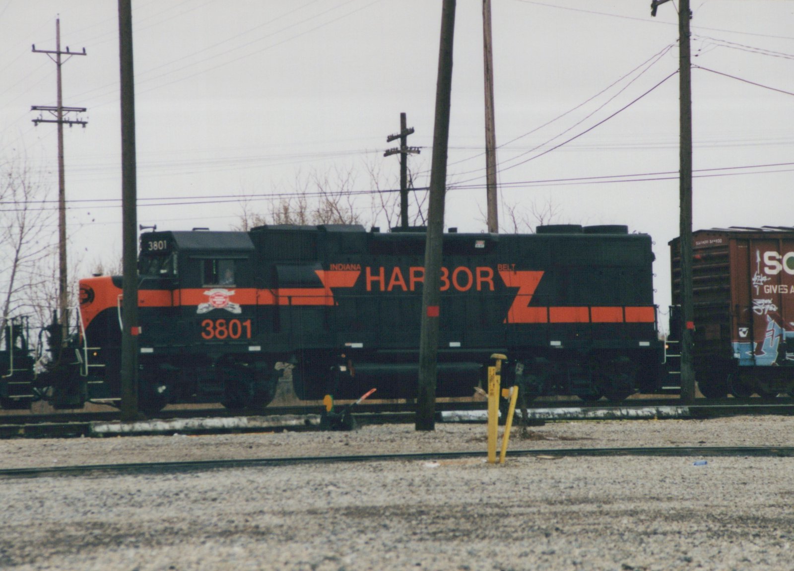 Black diesel locomotive with an orange nose and orange stripes, number 3801 and the word HARBOR on its side, on a track among utility poles, with a brown boxcar covered in graffiti at the right.