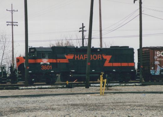 Black diesel locomotive with an orange nose and orange stripes, number 3801 and the word HARBOR on its side, on a track among utility poles, with a brown boxcar covered in graffiti at the right.