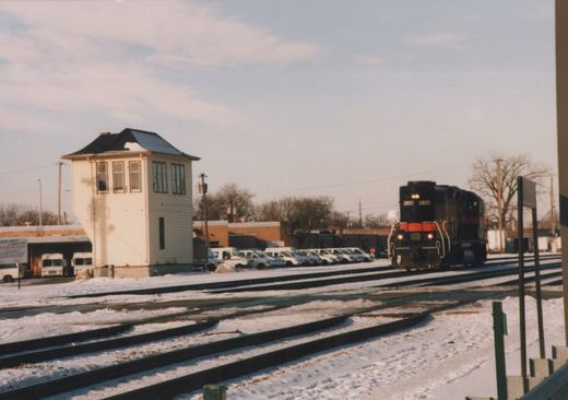 A black diesel locomotive with an orange nose band and the number 3801 on its number boards approaches on snowy tracks, beside a tall white wooden tower and a row of white postal vans.