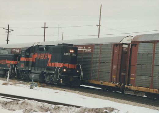 A black diesel locomotive with an orange stripe and HARBOR lettering, number 3802, headlights on, passes snowy ground beside a line of red and gray enclosed autorack cars.