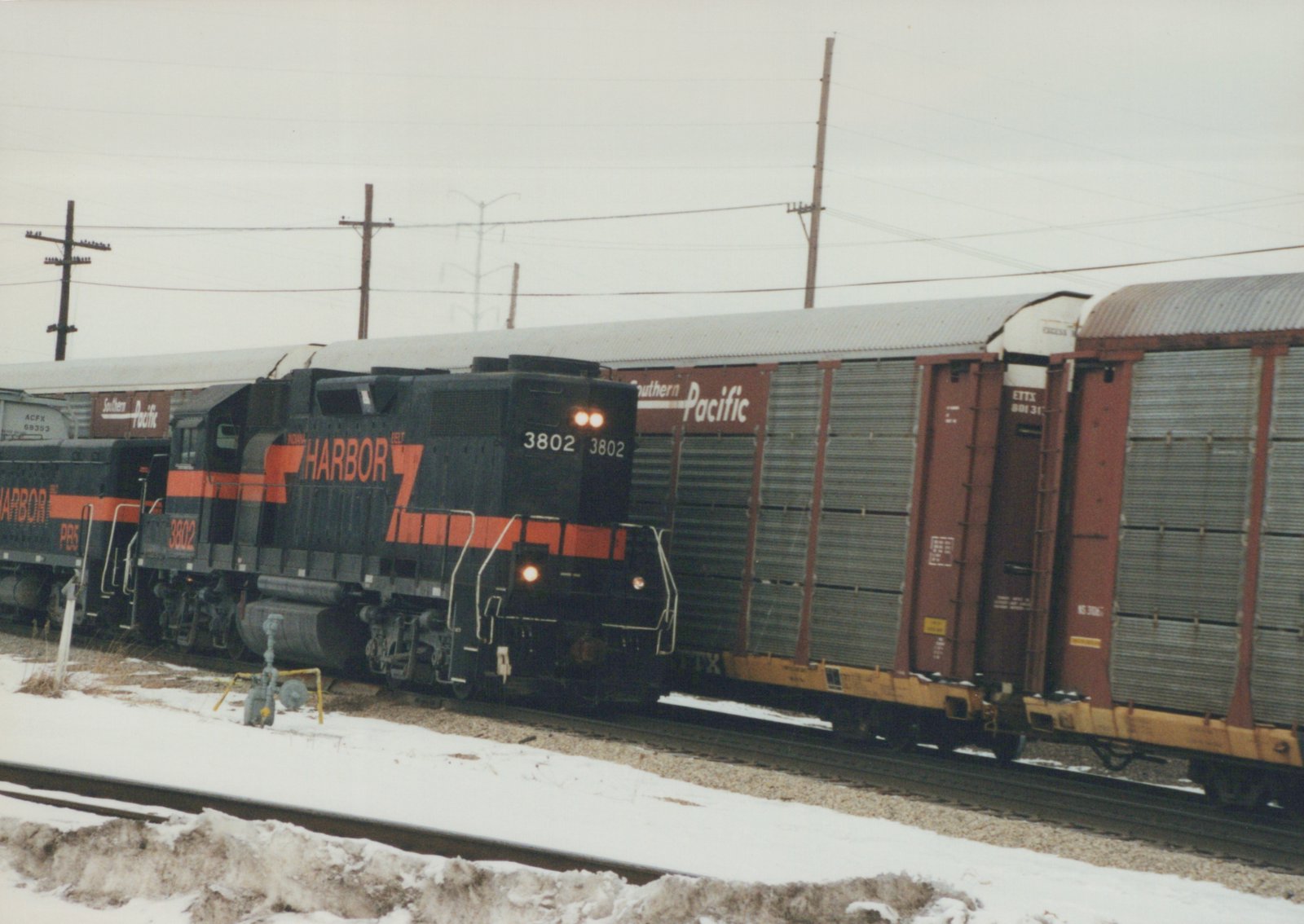 A black diesel locomotive with an orange stripe and HARBOR lettering, number 3802, headlights on, passes snowy ground beside a line of red and gray enclosed autorack cars.