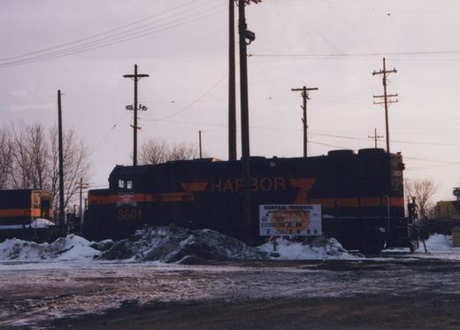 A black diesel locomotive with orange stripes and large orange HARBOR lettering, number 3801, stands behind piles of dirty snow and a white safety sign under utility poles and an overcast sky.