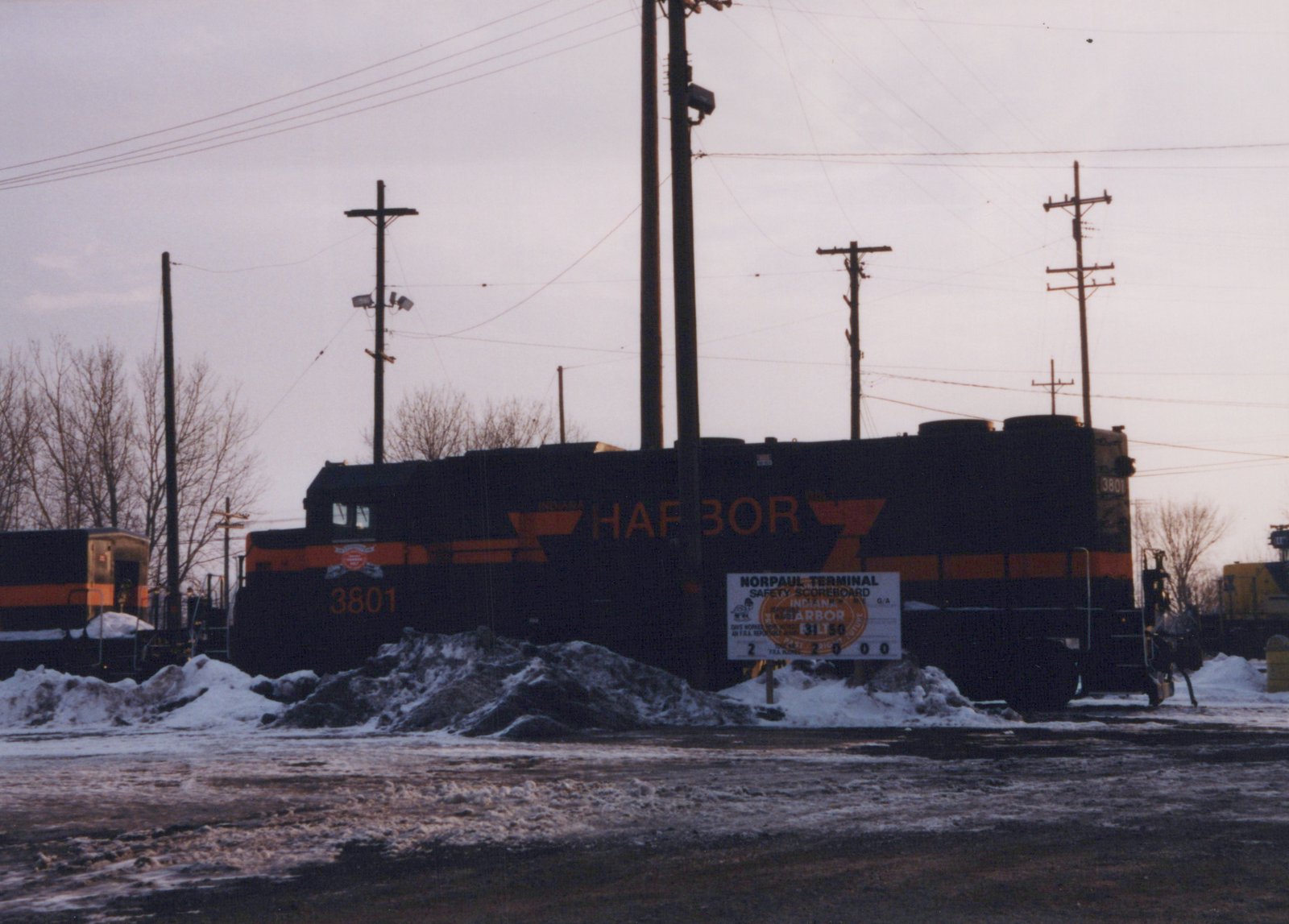 A black diesel locomotive with orange stripes and large orange HARBOR lettering, number 3801, stands behind piles of dirty snow and a white safety sign under utility poles and an overcast sky.