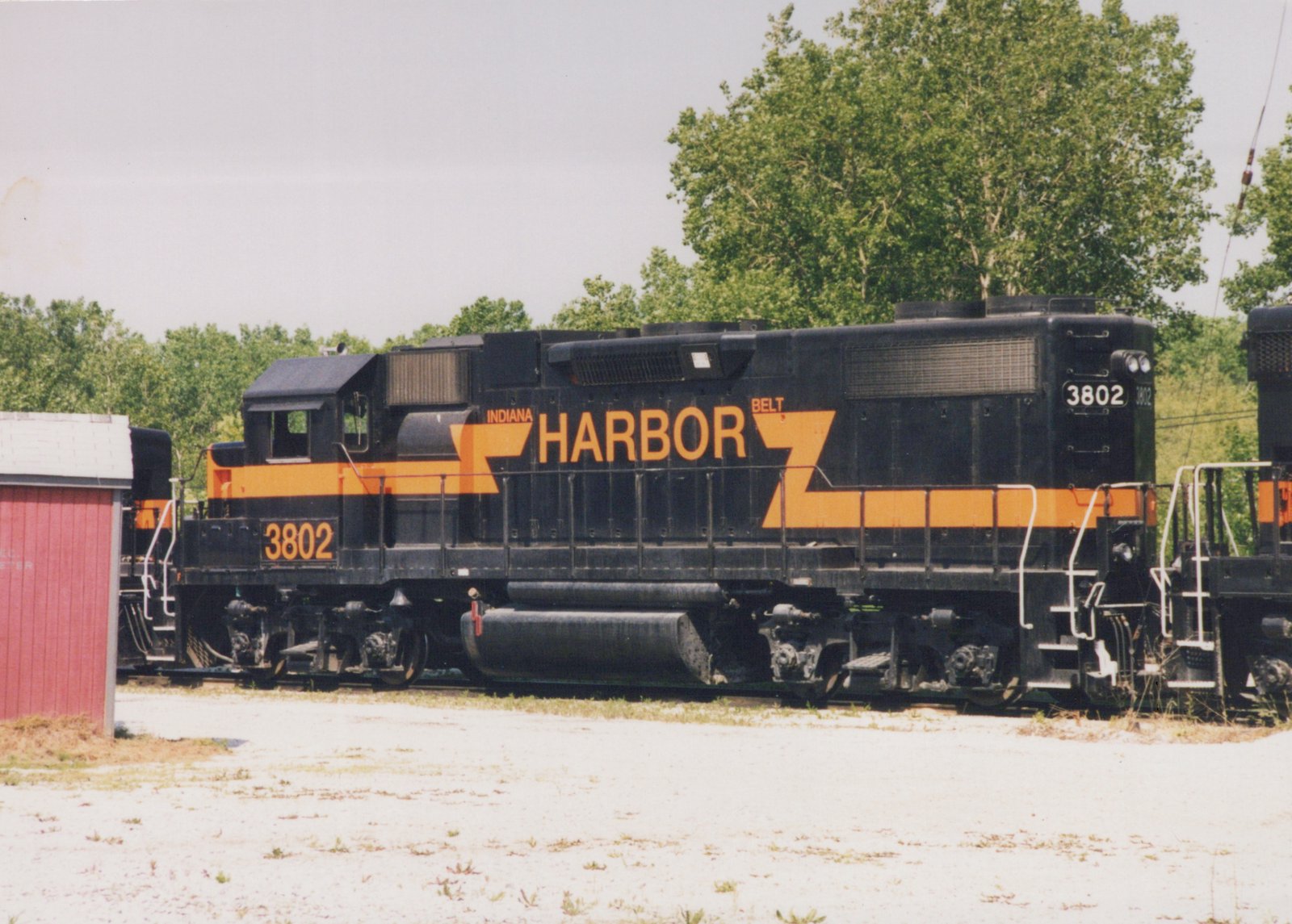 A black diesel locomotive with orange stripes and large HARBOR lettering, numbered 3802, seen from the side between two other locomotives, with trees behind and a red shed at the left.