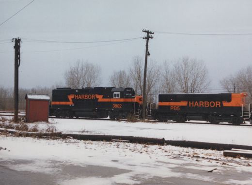 Two black and orange Indiana Harbor Belt locomotive units, 3802 and PB5, stand coupled on a snowy track beside a small red shed and utility poles under a gray sky.
