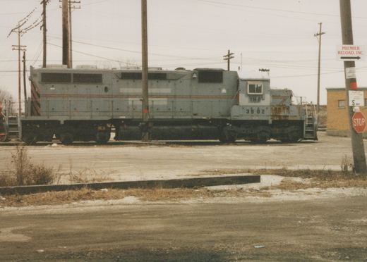 A gray six-axle diesel locomotive lettered IHB 3902 stands on a track beyond an empty gravel lot under an overcast sky, with utility poles and a pole carrying Premier Reload and stop signs at right.
