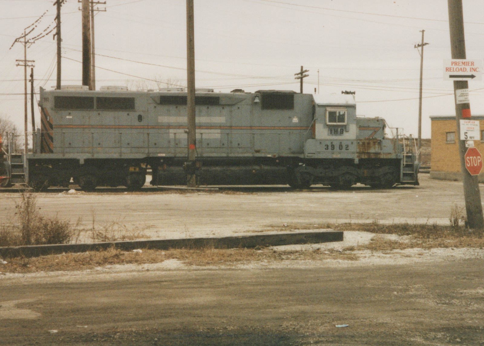A gray six-axle diesel locomotive lettered IHB 3902 stands on a track beyond an empty gravel lot under an overcast sky, with utility poles and a pole carrying Premier Reload and stop signs at right.