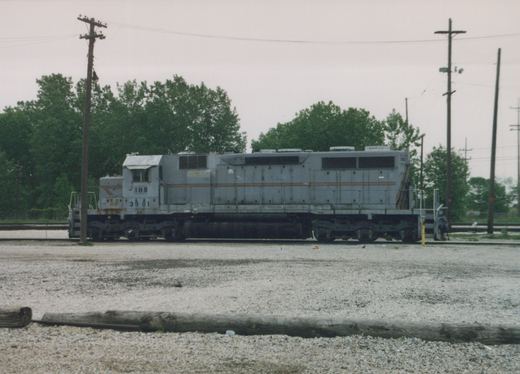 A gray six-axle diesel locomotive lettered IHB 3901 is shown from the side on yard track, with trees and utility poles behind it and an old wooden tie in the foreground.