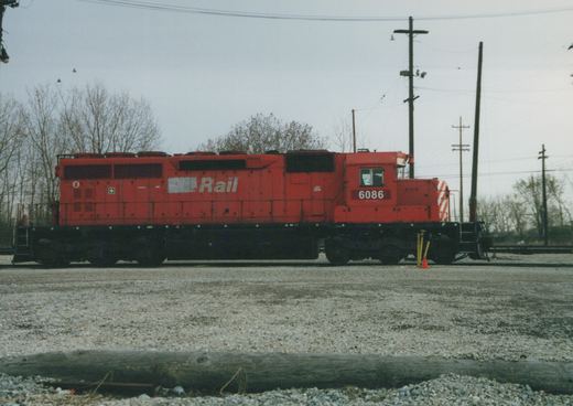 Side view of a red six-axle diesel locomotive numbered 6086, lettered 'Rail' beside a white patch, standing on a track behind a gravel lot under an overcast sky with bare trees and utility poles.